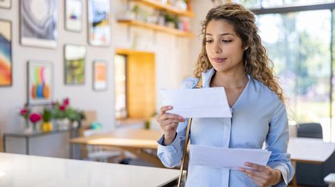 Une jeune femme blonde aux cheveux bouclés lit une lettre qu'elle vient de recevoir, debout dans son salon
