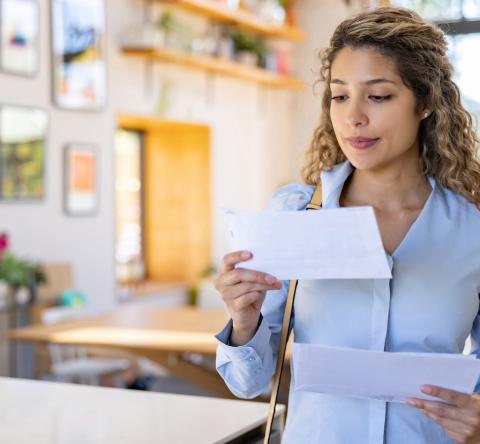 Une jeune femme blonde aux cheveux bouclés lit une lettre qu'elle vient de recevoir, debout dans son salon